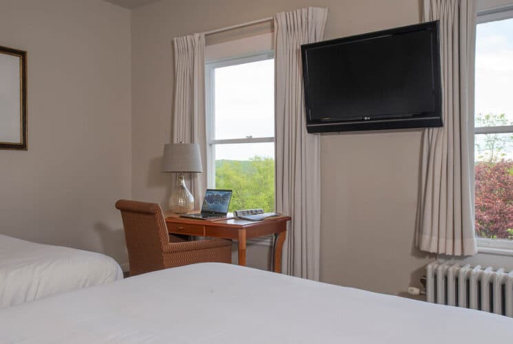 Bedroom with two double beds in white linens, writing desk with lamp and computer by a large window with curtains and TV on the wall