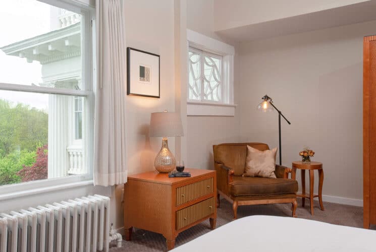 Bedroom with a double bed, dresser with lamp, leather sitting chair with lamp and side table and large window with white curtains