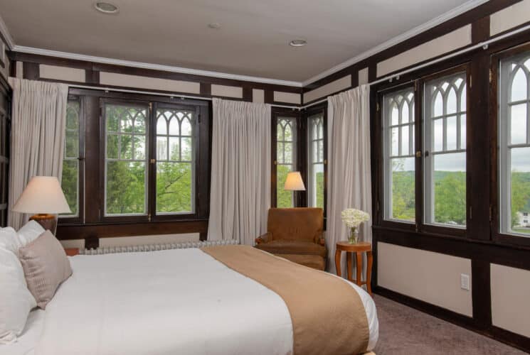 Bedroom with walls of windows with dark wood detail, queen bed and sitting chair in the corner with side table and lamp