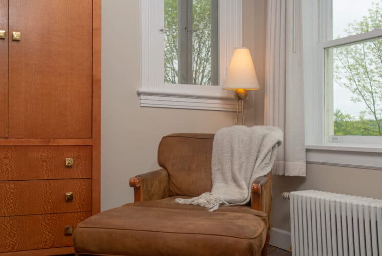 Corner of a bedroom with a plush sitting chair and ottoman, tall armoire and window with white curtains