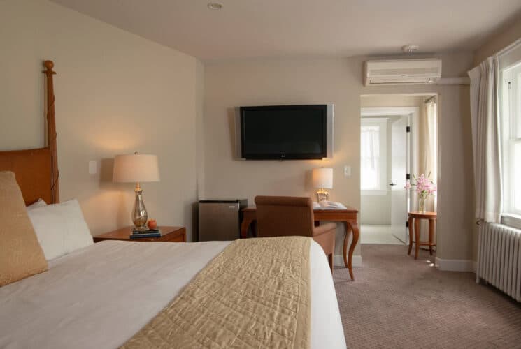 Bedroom with king bed in white linens, writing desk under a TV on the wall, mini fridge and doorway into an attached bathroom