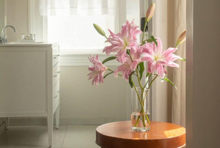 A small side table with a vase of pink lilies in a bathroom with white tile and a white sink vanity by a window