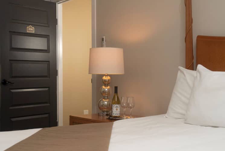 Bedroom with a black door, queen bed in white linens and side table with lamp and bottle of wine with two glasses.