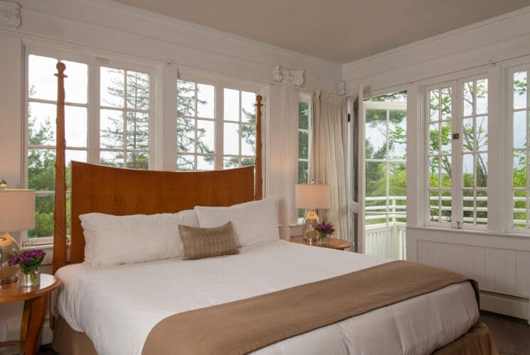 Bright bedroom with walls of windows, king bed, side tables with lamps nd flowers and doorway open to an outdoor patio