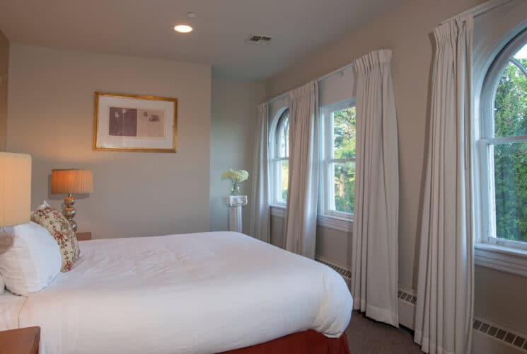 Elegant bedroom with a queen bed in white linens, side tables with lamps and arched windows with white curtains