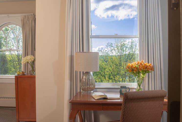 A writing desk with chair, lamp, book, vase of flowers and coffee mug in front of a large window in a bedroom