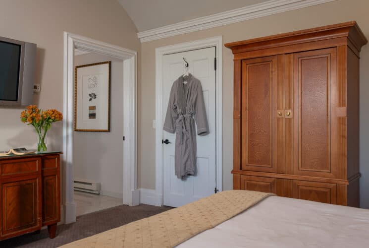 Bedroom with queen bed, tall armoire, dresser with TV and plush robe hanging on the back of the door