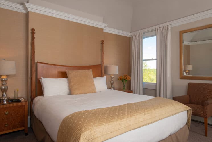 Bedroom with king bed in white and gold linens, side tables with silver lamps and window with white curtains