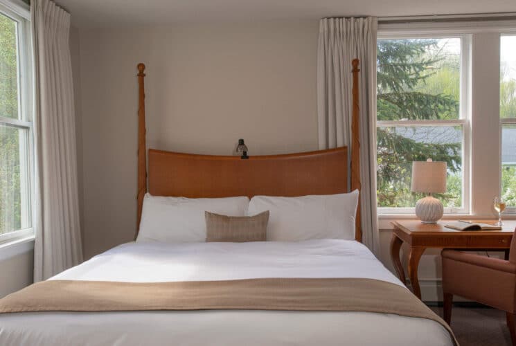 Bedroom with queen bed in white and tan, writing desk with lamp, book and glass of wine by a window with white curtains