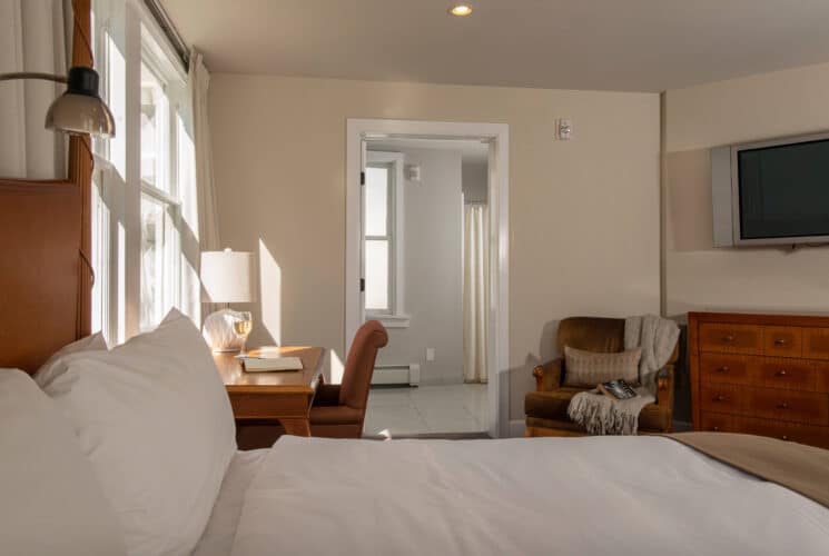 Bedroom with queen bed in white linens, writing desk, plush sitting chair and dresser with TV