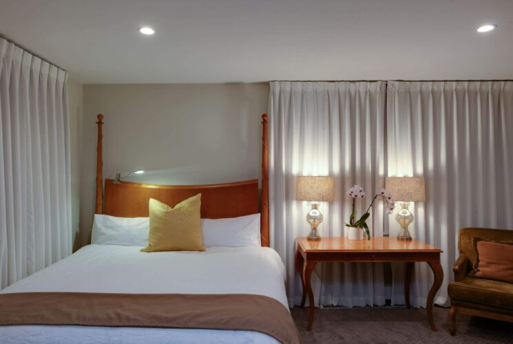 Bedroom with queen bed in white with a tan blanket and yellow pilloww, table with two lamps and white curtains all around