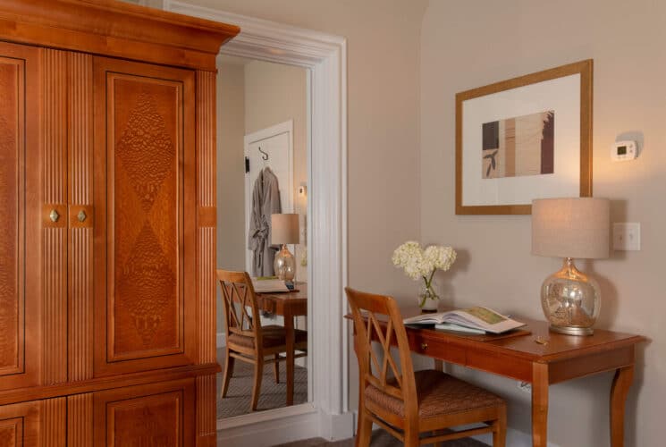 Bedroom with writing desk, chair, book, lamp and vase of flowers next to a large mirror and tall armoire cabinet