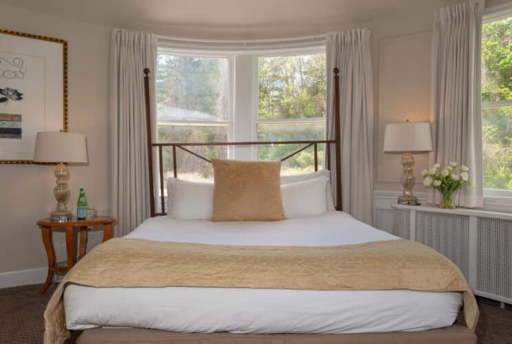 Bedroom with queen bed in front of a large window with white cuurtains, side table with lamp and vase of white flowers