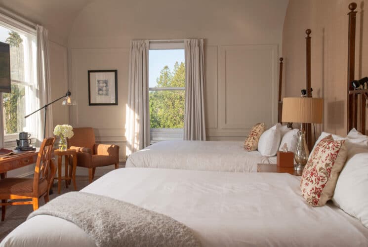 Bright and spacious bedroom with two double beds, writing desk with chair and sitting chair by two large windows with white curtains