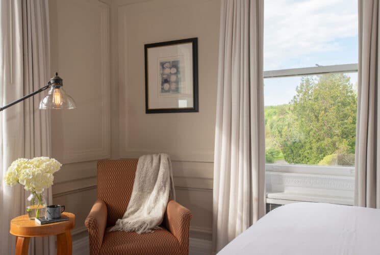 Corner of a bedroom with a sitting chair, table with vase of flowers and coffee mug, lamp by large windows with white curtains