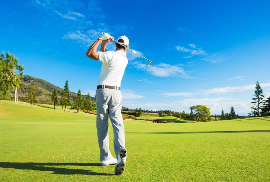 A man in grey pants and white shirt mid swing on a golfing green with blue skies above