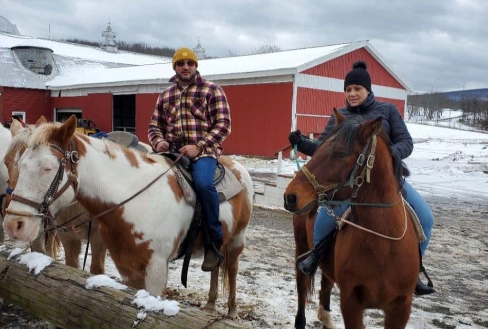 Two people on different colored horses at a farm with a large red barn