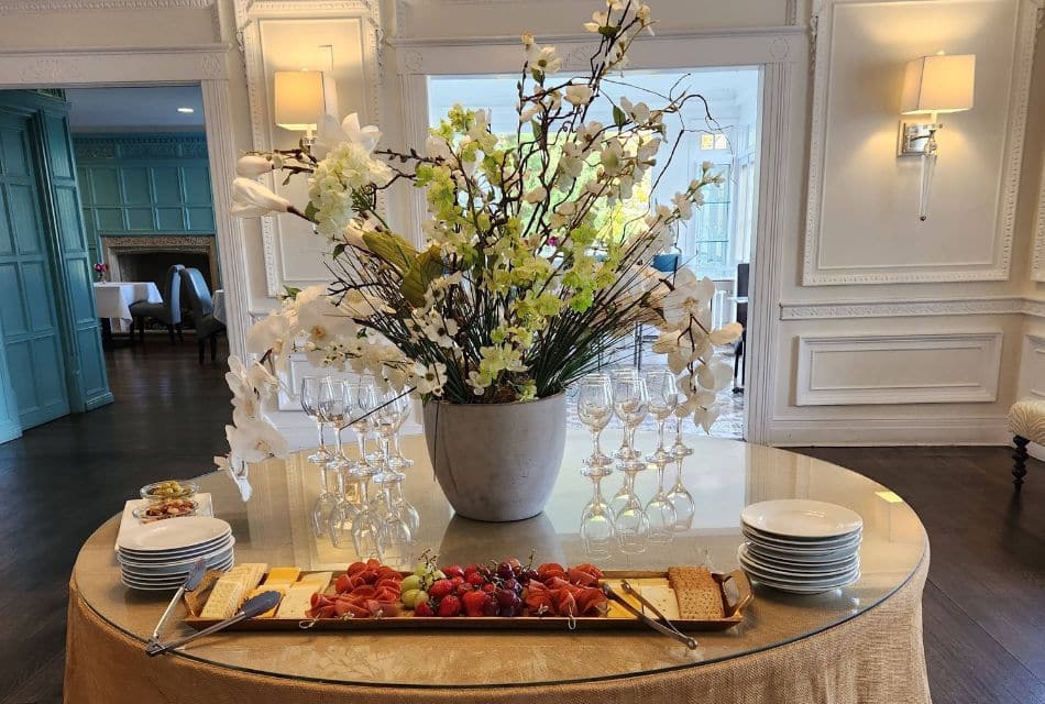 A round glass topped table with gold linens holding a large vase of flowers, charcuterie platter, appetizer plates and clear wine glasses