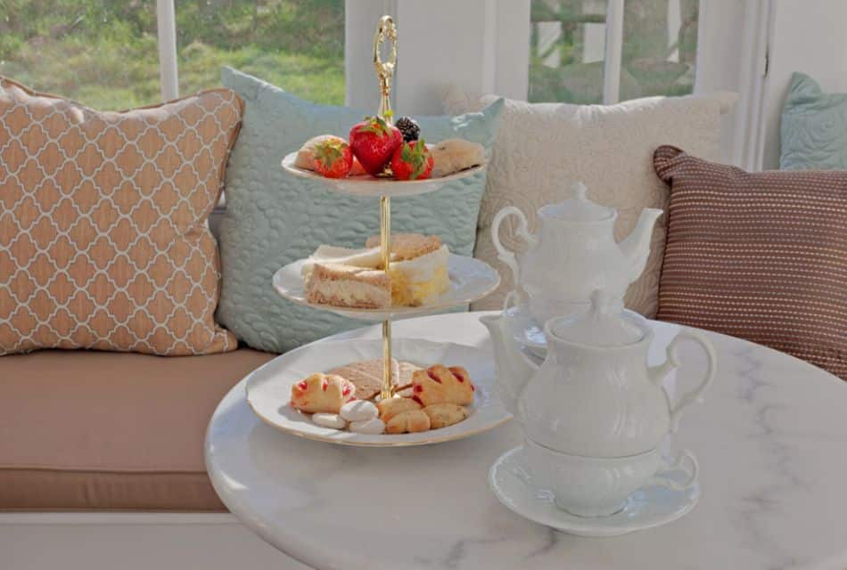 A three-tiered stand with various desserts and fruits on a round marble table with two white tea pots