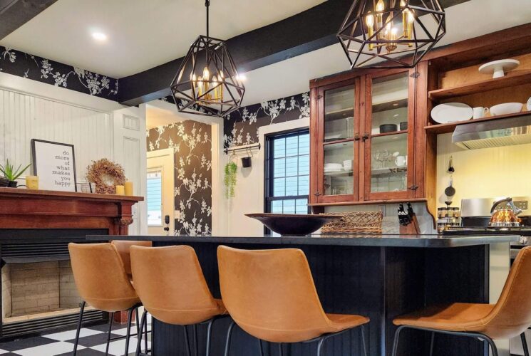 Kitchen with black and white floral wallpaper, island with leather bar stools and fireplace with mantle