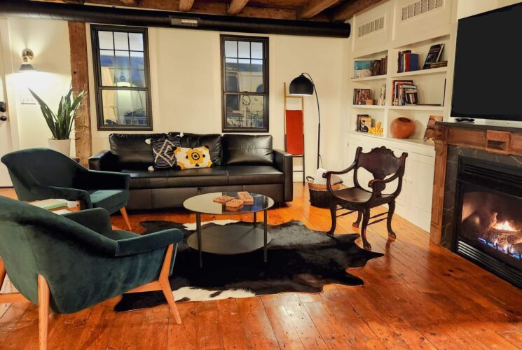 Living room with hardwood floors, black leather couch, velvet sitting chairs and fireplace with TV above