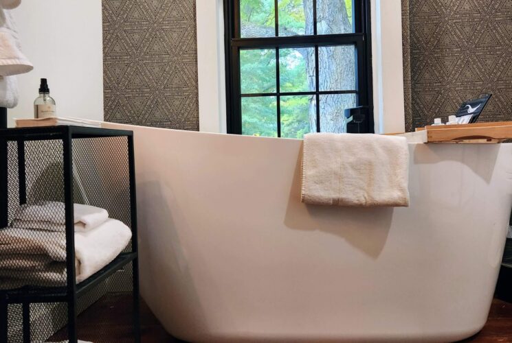 Large white modern soaking tub under a window next to shelving with folded white towels