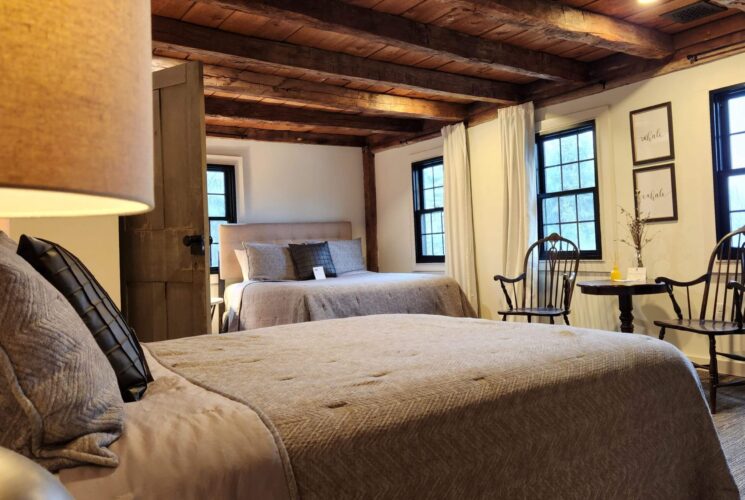 A spacious bedroom suite with two queen beds, rustic wood beam ceiling, table and two charis and several windows with white curtains