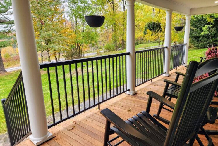 An expansive front porch of a home with a black railing and black rocking chairs overlooking a lawn and trees