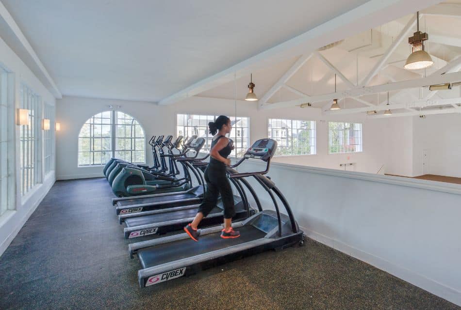 A woman running on a treadmill that is next to other treadmills and ellipticals on an upper loft in a pool house
