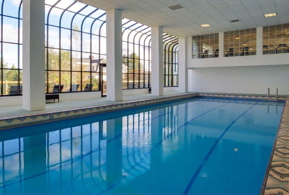 A large indoor pool with a wall of tall windows and an upper loft with treadmills