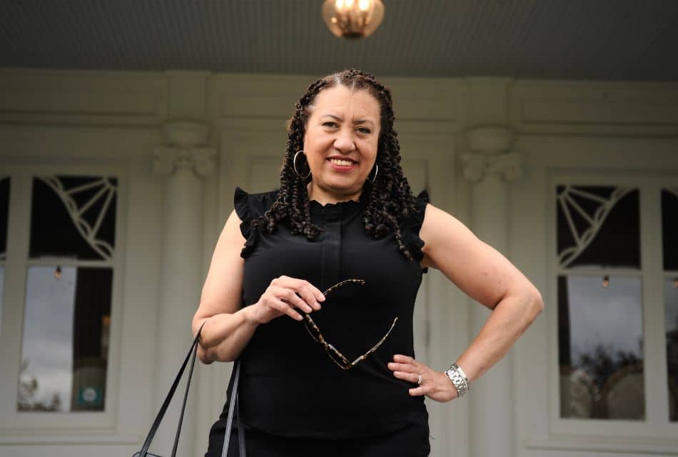 A beautiful woman in a black sleeveless dress holding a purse and sunglasses, standing in front of a white home