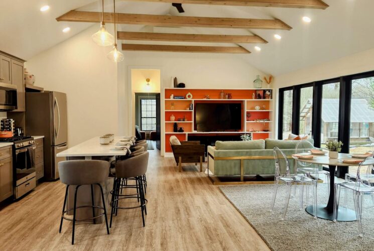 Open concept kitchen and living room of a home with island and bar stools, dining table with four chairs, couch and chairs in front of TV with built in shelving