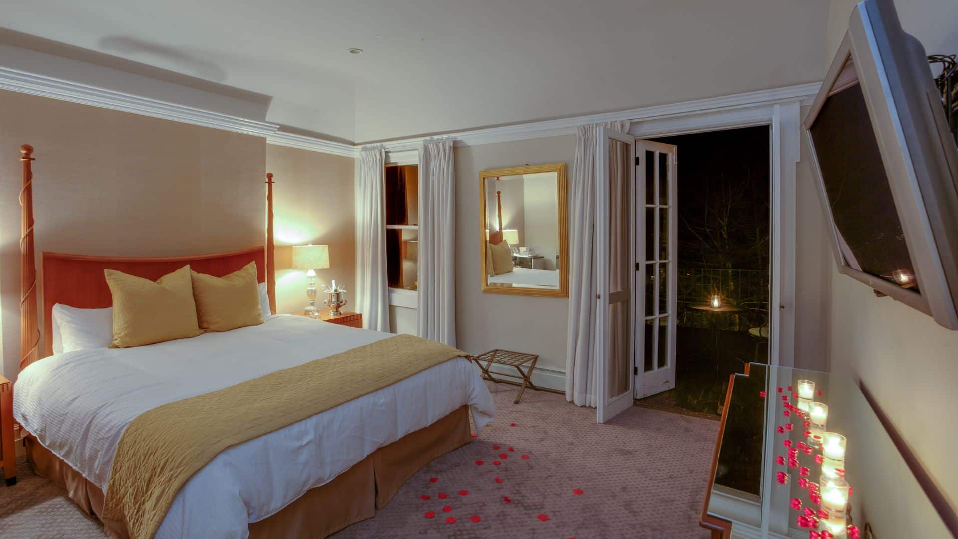 A beautiful bedroom with queen bed in gold and white linens, candles lit on a mirror topped dresser and flower petals strewn on the floor