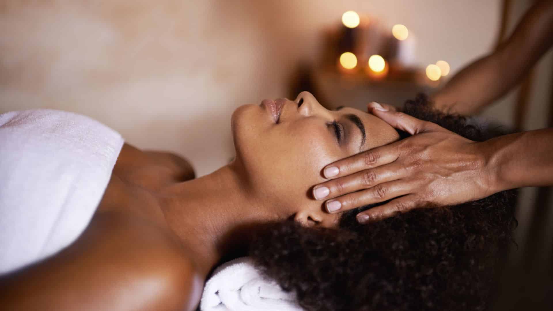 A woman laying down on a massage table with a white towel over her chest and a second persons hands on her head