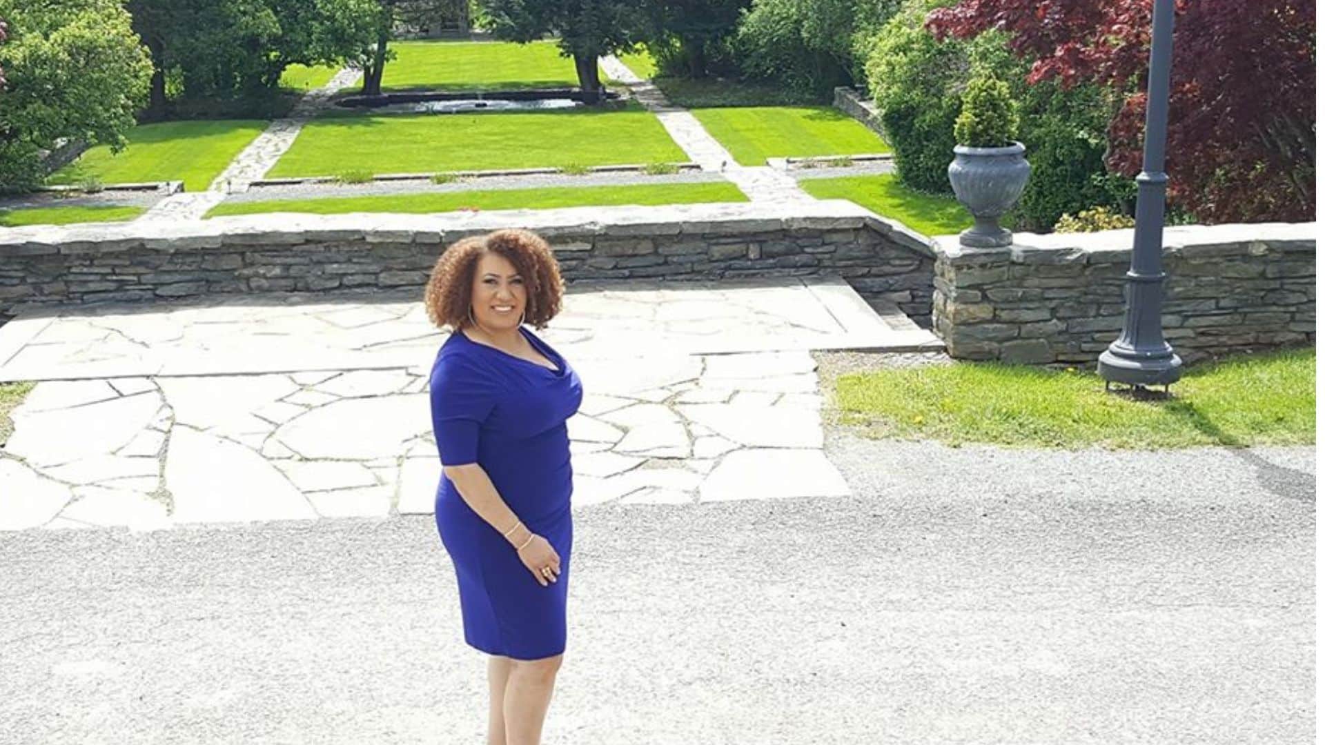 A beautiful woman in a blue dress standing on an outdoor patio with an expansive manicured lawn and tall trees behind her