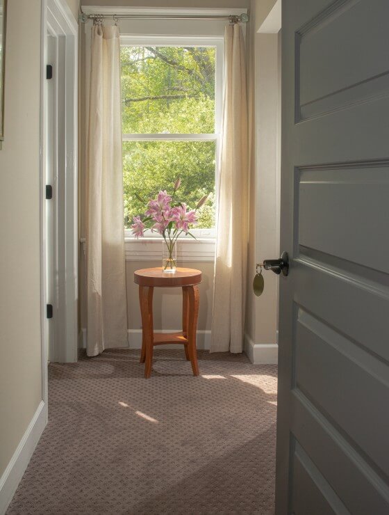 An open door showing a single brown table with a vase of pink flowers under a window with cream curtains