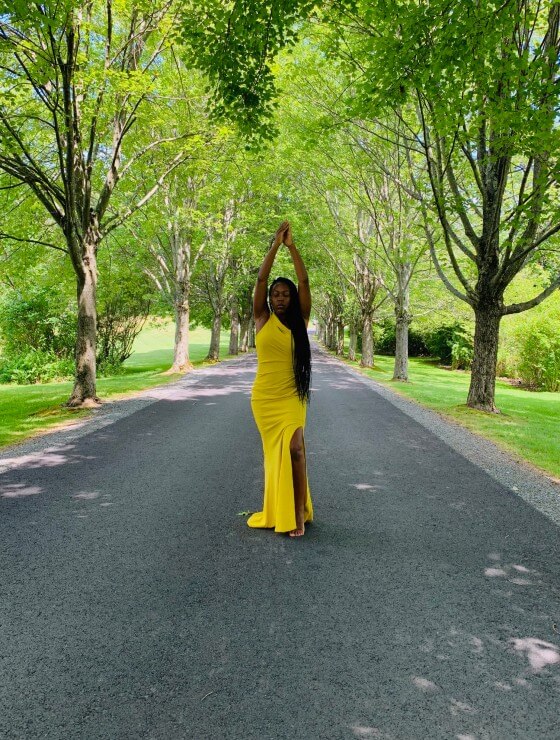 A woman in a yellow dress posing on an empty road lined with tall green trees