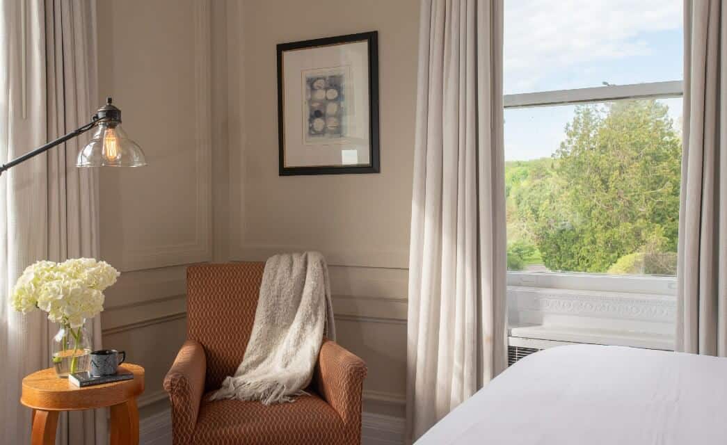 Corner of an elegant bedroom with an upholstered chair, table with coffee cup and vase of flowers and large window with white curtains