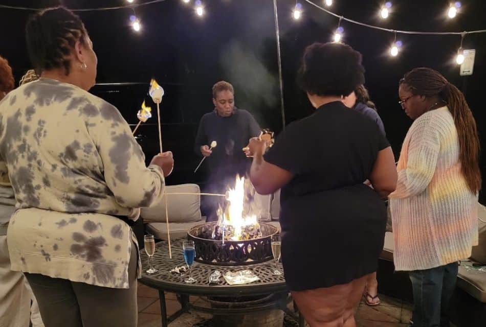 A group of women of various ages standing around an outdoor fire pit roasting marshmallows