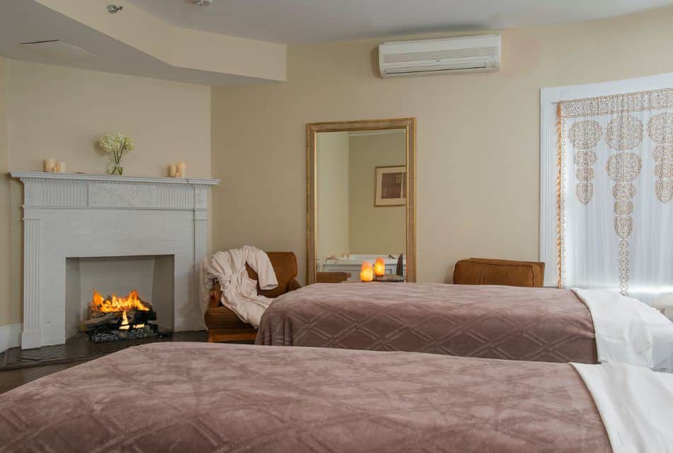Soft and cozy room with two massage tables covered in blankets, fire going in a fireplace and sitting chairs with a plush robe