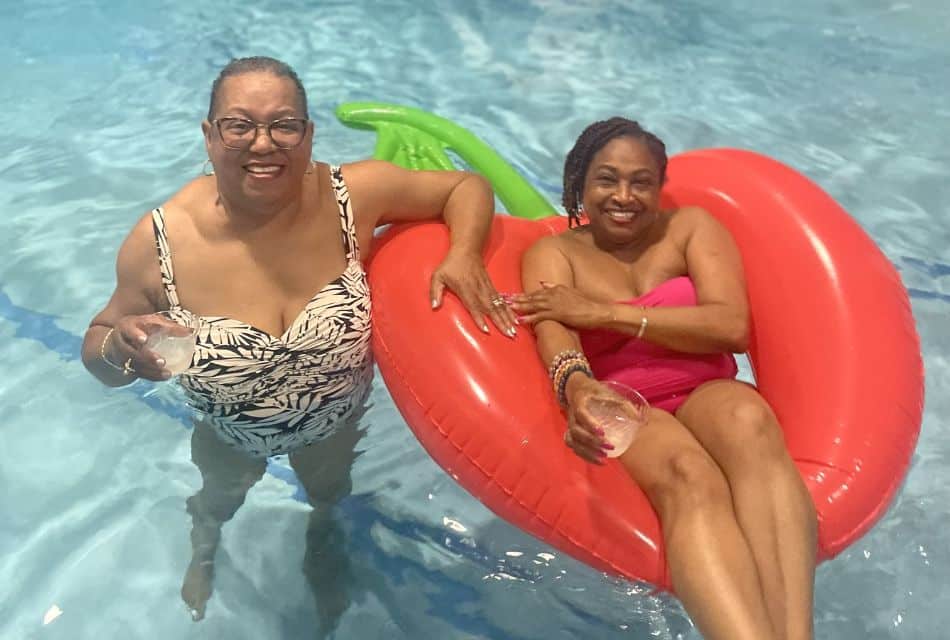 Two women in a pool, one holding a drink and the other floating in a red blow up pool float