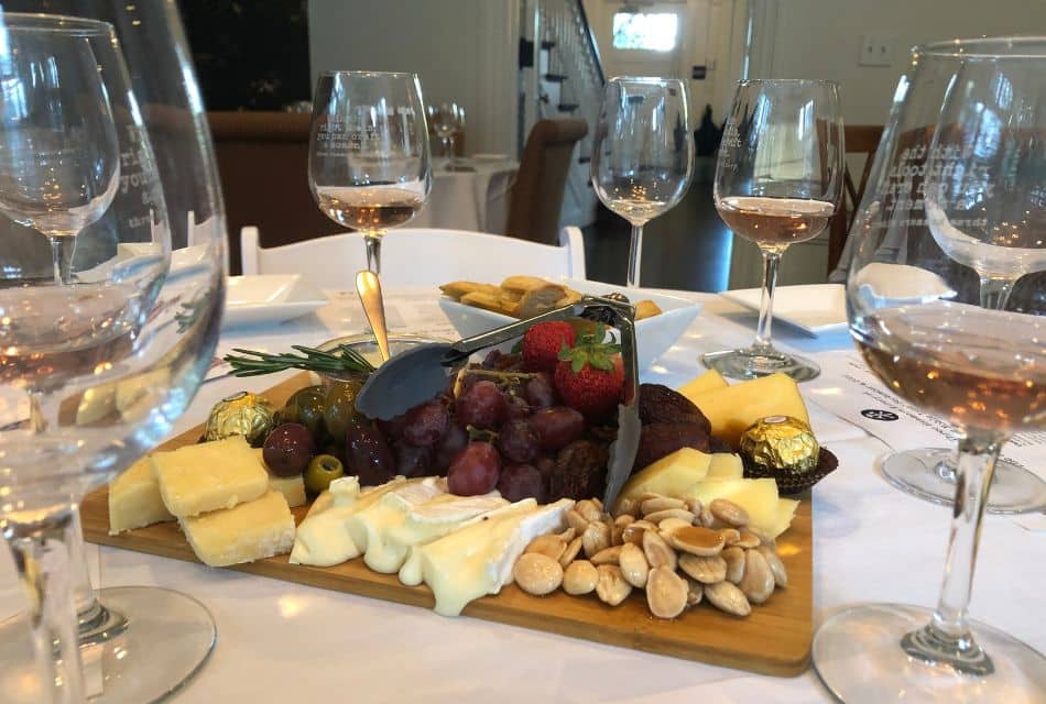 A wood charcuterie platter with cheeses, grapes, olives and crackers on a white table with wine glasses