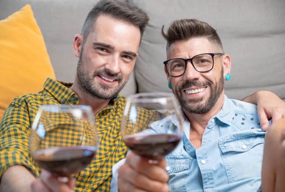 Two men sitting together in front of a couch, each holding up a glass of red wine