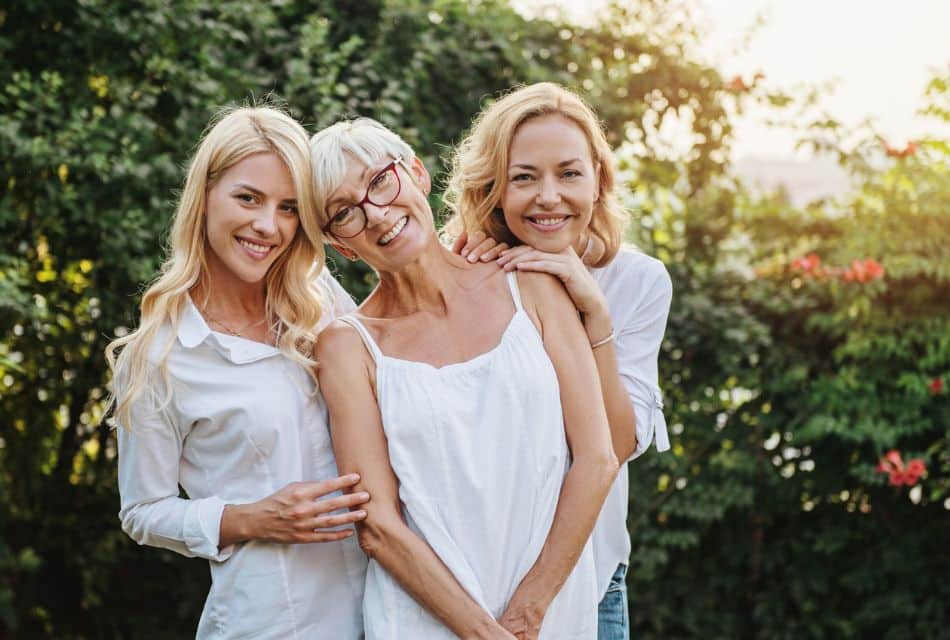 Three generations of women, each wearing white standing together outside by trees bathed in sunshine