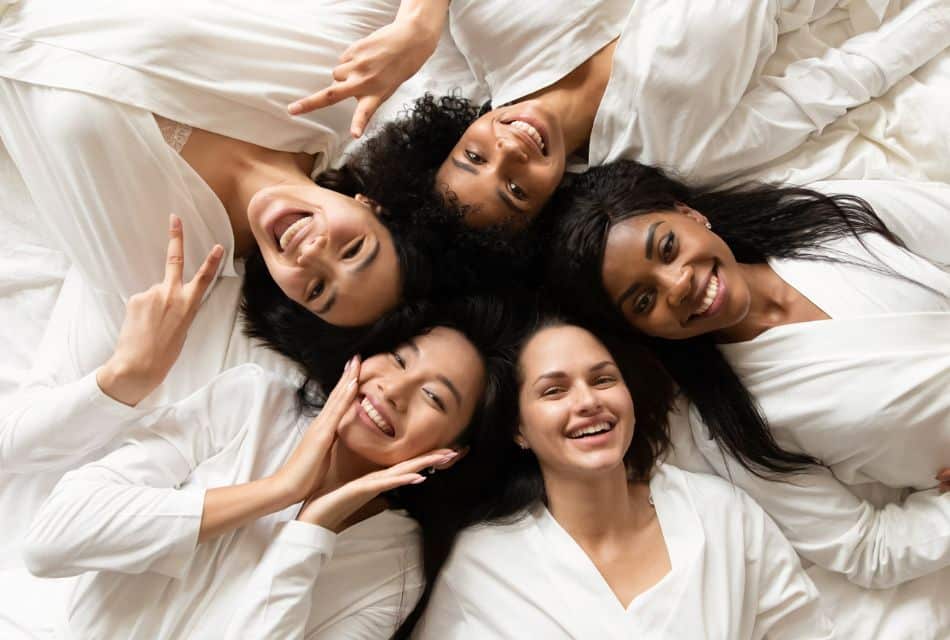 Five women, each wearing a white robe, laying in a circle with their heads together