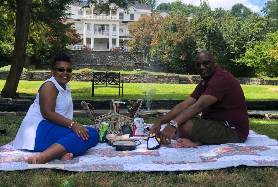 A couple sitting on a quilt outside with a picnic basket and food, on a lawn in front of a large stately home