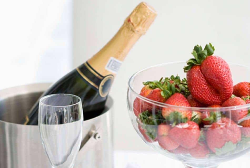 A bowl of strawberries next to a wine glass and silver bucket with a bottle of champagne