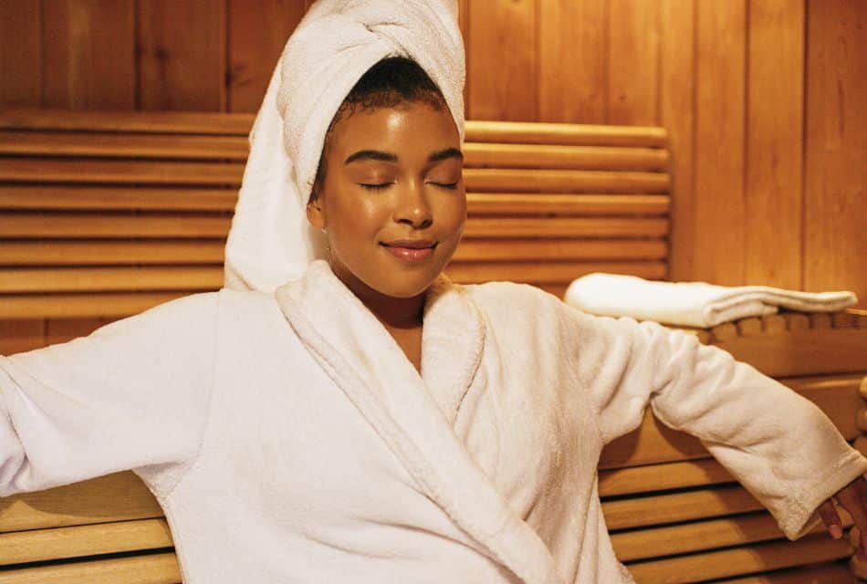 A woman in a plush white robe and towel on her head sitting on a bench in a sauna