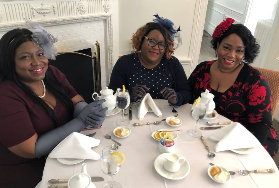 Three ladies in colorful dresses and hats sitting at a white table set for tea time