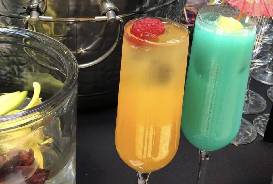 Two fruity cocktails, one orange and one green on a table with other glasses and a silver bucket